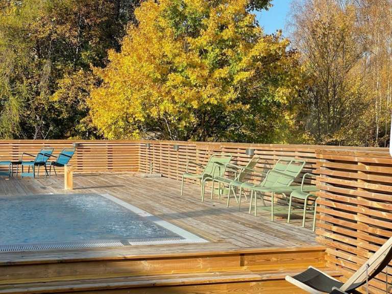 Ein türkisfarbener Whirlpool ist von einer Holzterrasse mit vier grünen Liegestühlen und einem Baum im Herbstlaub umgeben.
