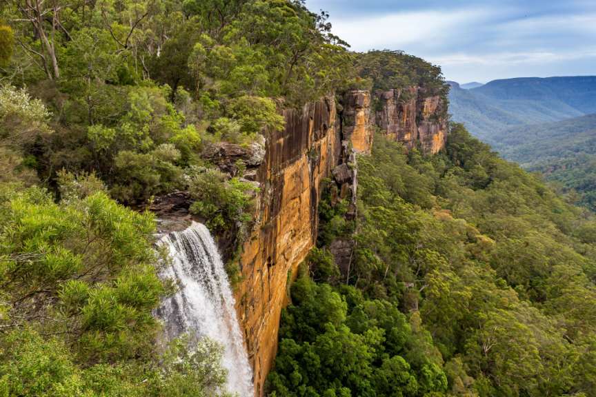 Ein Wasserfall stürzt von einer orangefarbenen Felsklippe, die von üppigem grünen Wald umgeben ist, in ein bergiges Tal.