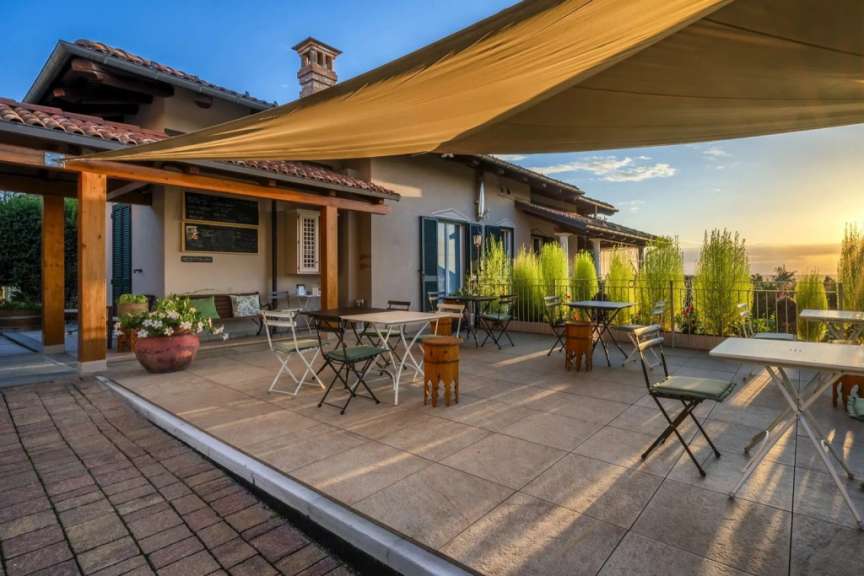 Ein sonnenbeschienener Terrassenbereich mit Tischen und Stühlen unter einer Sonnensegelkonstruktion vor einem Gebäude.