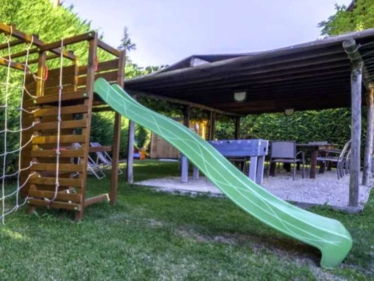 Ein Spielturm mit Rutsche steht auf einem Rasen vor einer überdachten Terrasse mit Tisch und Stühlen.