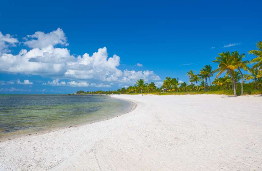 Ein ruhiger Strand mit weißem Sand, klarem türkisfarbenem Wasser und Palmen unter einem strahlend blauen Himmel mit weißen Wolken.