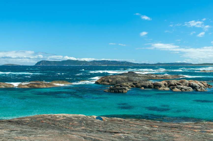 Felsformationen ragen aus dem türkisfarbenen Wasser einer Bucht, während im Hintergrund eine bewaldete Küstenlinie unter einem blauen Himmel mit Wolken zu sehen ist.