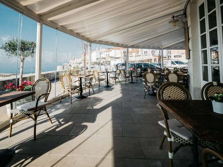 Ein schattiger Außenbereich eines Restaurants mit Tischen und Stühlen, die am Meer und an einer Straße liegen.