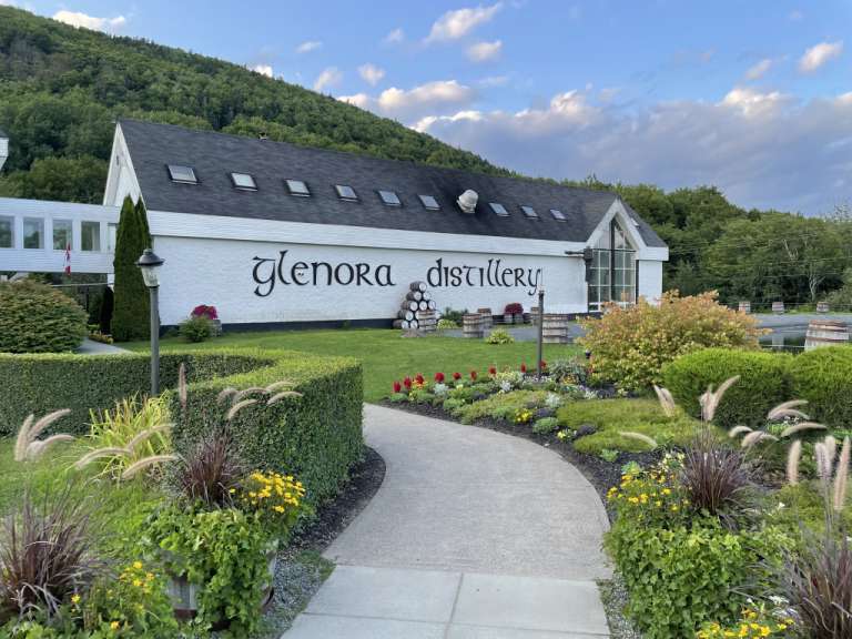 Ein Gehweg schlängelt sich durch einen Garten vor der Glenora Distillery, die von einer grünen Hügelseite und Bäumen umgeben ist.