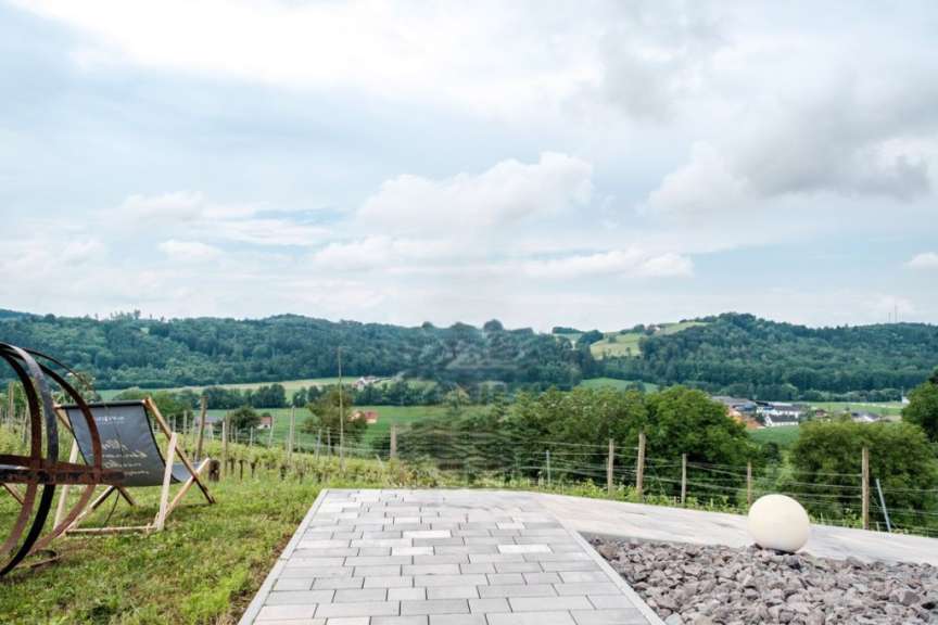 Eine Aussicht von einer Terrasse mit Rasen, einer Liege, einem Weg aus grauen Pflastersteinen und einem Kiesbeet mit einer weißen Kugel, vor einer hügeligen, bewaldeten Landschaft unter einem bewölkten Himmel.