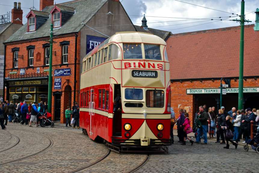 Eine rote und cremefarbene historische Straßenbahn fährt auf einem Kopfsteinpflasterweg durch eine viktorianische Straße, vorbei an Geschäften und einer Menge von Menschen.