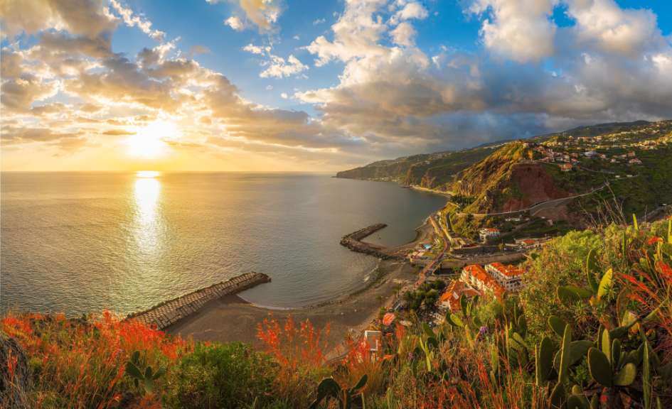 Ein Panorama zeigt die Küste mit dem Meer und dem Sonnenuntergang, eine Ortschaft an den Hängen und Kakteen im Vordergrund.