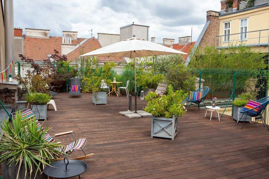 Eine geräumige Dachterrasse mit Holzbelag, verschiedenen Sitzgelegenheiten, Pflanzen und einem großen Sonnenschirm ist bei bewölktem Himmel zu sehen.