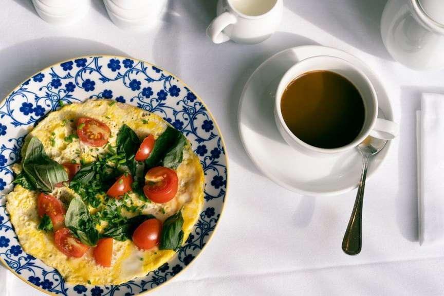Ein Omelett mit Tomaten und Kräutern liegt auf einem blauen und weißen Teller neben einer Tasse Kaffee auf einer weißen Tischdecke.