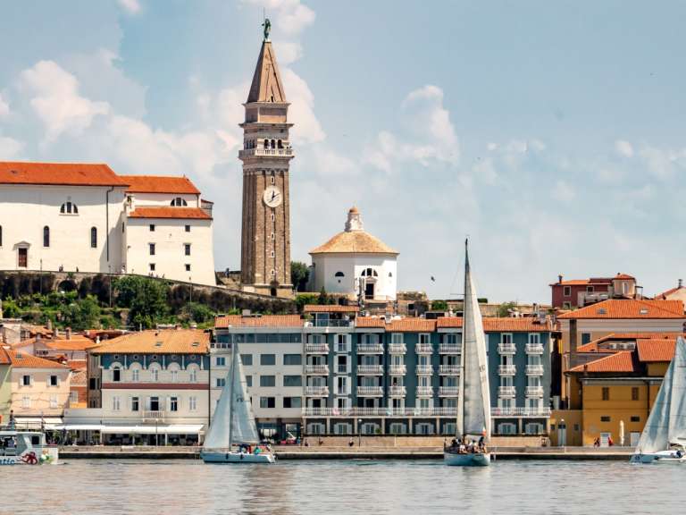 Ein Sommerpanorama mit segelnden Booten vor einer Küstenstadt mit traditionellen Gebäuden und einem Kirchturm.