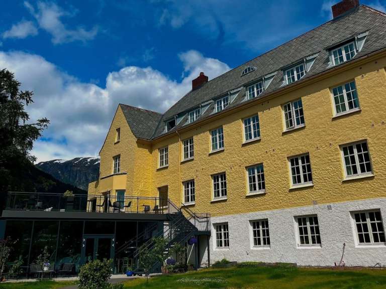 Ein gelbes Gebäude mit weißen Fenstern steht auf einer grünen Wiese unter einem blauen Himmel mit Wolken.