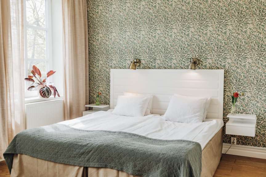 Ein Doppelbett mit weißer Bettwäsche und einer grünen Decke steht vor einer grünen Tapete mit Blattmuster, neben einem Fenster mit hellrosa Vorhängen.