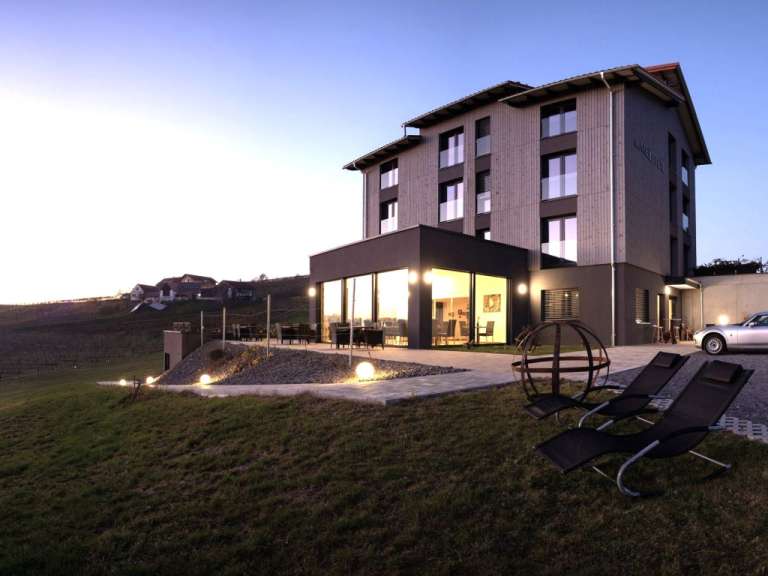 Ein modernes Hotelgebäude mit großen Fenstern und einem angrenzenden Restaurantbereich steht auf einem Hügel neben einem Weingut, während zwei Liegestühle auf einem Rasen davor stehen und die Sonne untergeht.