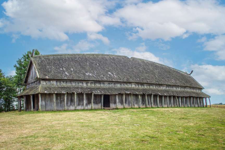 Ein großes, altes Wikingerhaus mit Holzdach steht auf einer grünen Wiese unter einem bewölkten Himmel.