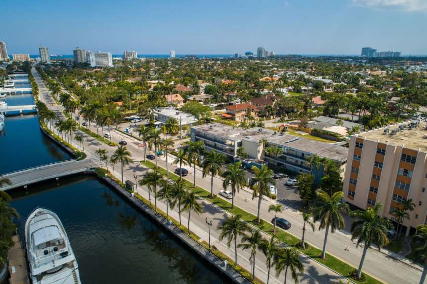 Eine Drohnenaufnahme zeigt eine breite Straße mit Palmen, die von einem Kanal mit einer großen Yacht und mehreren Gebäuden mit Blick auf den Ozean gesäumt ist.