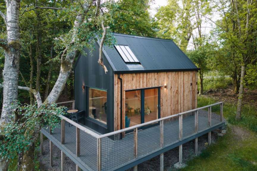 Ein modernes, hölzernes Ferienhaus mit Dachfenstern und einer umlaufenden Veranda steht inmitten eines üppigen, grünen Waldes.