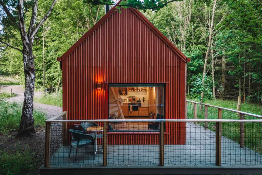 Ein kleines rotes Haus mit Wellblechfassade steht in einem Wald, davor eine Veranda mit zwei Stühlen und einem Tisch.