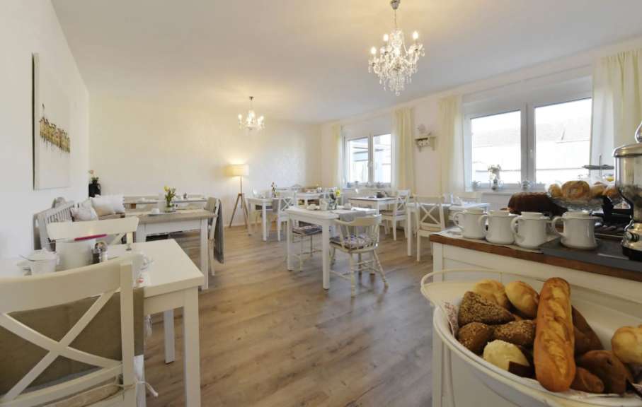 In einem hellen Frühstücksraum stehen mehrere Tische und Stühle, an einem Buffet werden verschiedene Brotsorten präsentiert.