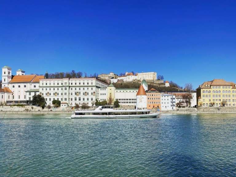 Ein Boot fährt auf einem Fluss an einer Stadt mit verschiedenen Gebäuden und einem Schloss im Hintergrund vorbei.