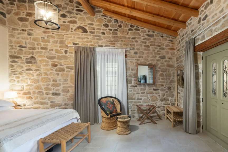 In einem Zimmer mit Steinwänden und Holzbalkendecke sind ein Bett, ein Sessel und eine Tür zu sehen.