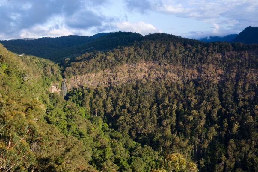 Dichte Wälder bedecken Hügel und Täler, während eine schmale Wasserfall auf einer felsigen Klippe herabstürzt.