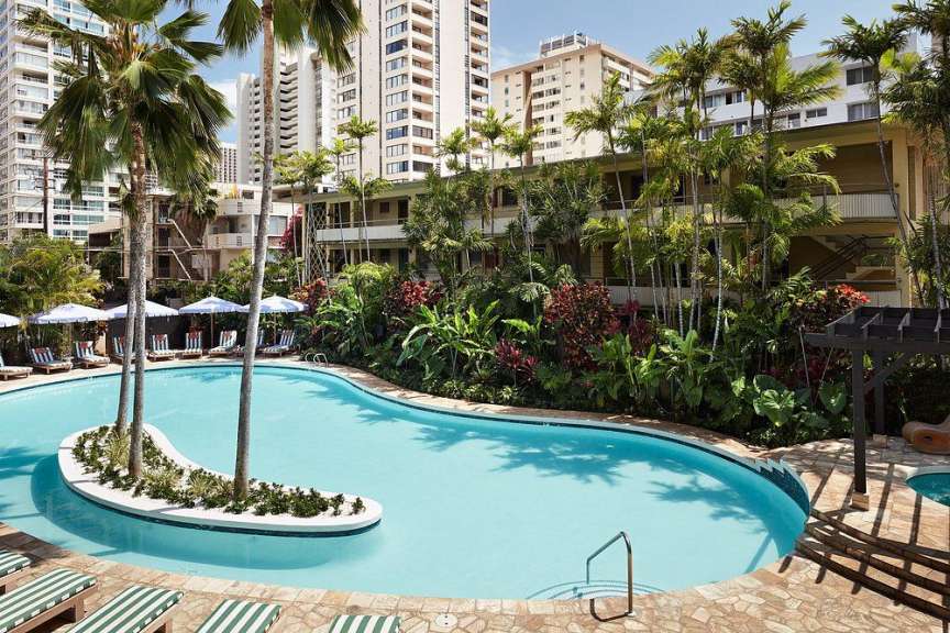 Eine geschwungene Poollandschaft mit Palmen und Liegestühlen vor einem Hotelgebäude.