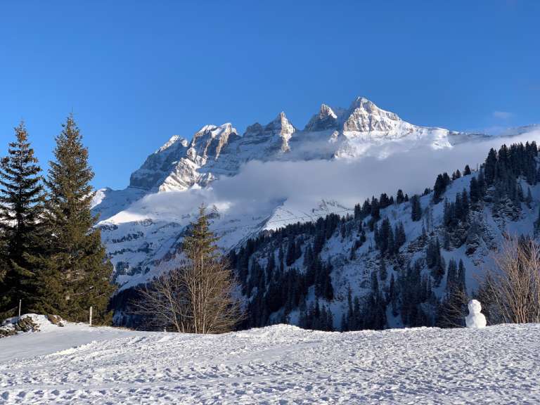 Eine verschneite Berglandschaft unter klarem blauem Himmel mit schneebedeckten Gipfeln und einem kleinen Schneemann im Vordergrund.
