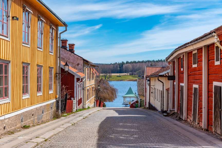 Eine kopfsteingepflasterte Straße führt zu einem See, gesäumt von bunten Holzhäusern unter einem strahlend blauen Himmel mit weißen Wolken.