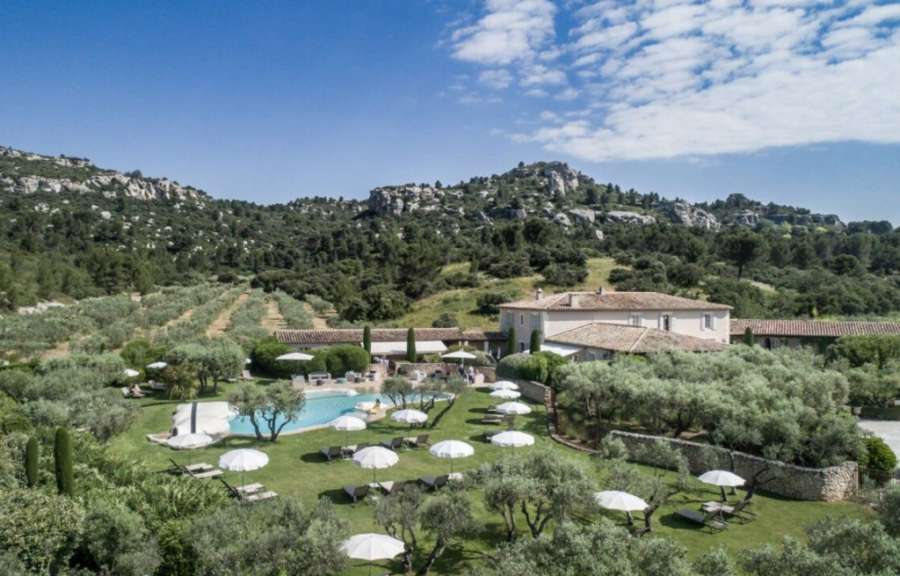Ein luxuriöser Poolbereich mit vielen Sonnenschirmen und Liegestühlen ist von Olivenbäumen und einem großen Gebäude im Hintergrund umgeben, das vor einer hügeligen, baumbestandenen Landschaft mit felsigen Gipfeln liegt.