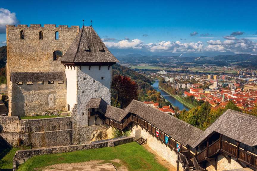Dieses Bild zeigt eine Ansicht von oben auf eine Burg mit einem roten Ziegeldach und einer Stadtlandschaft im Hintergrund unter einem blauen Himmel mit Wolken.