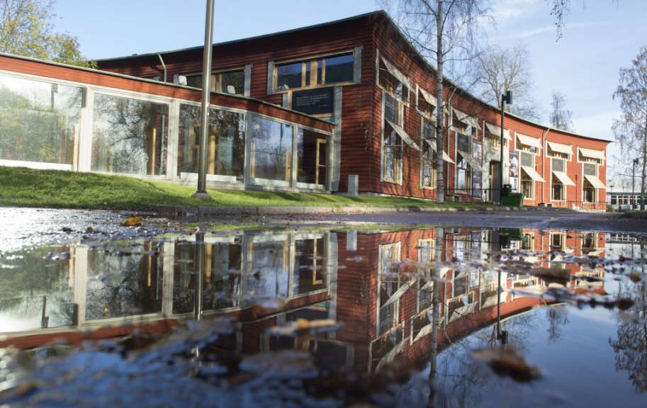 Ein rotes Gebäude mit vielen Fenstern spiegelt sich in einer Pfütze auf dem Boden, in der sich auch Blätter befinden.