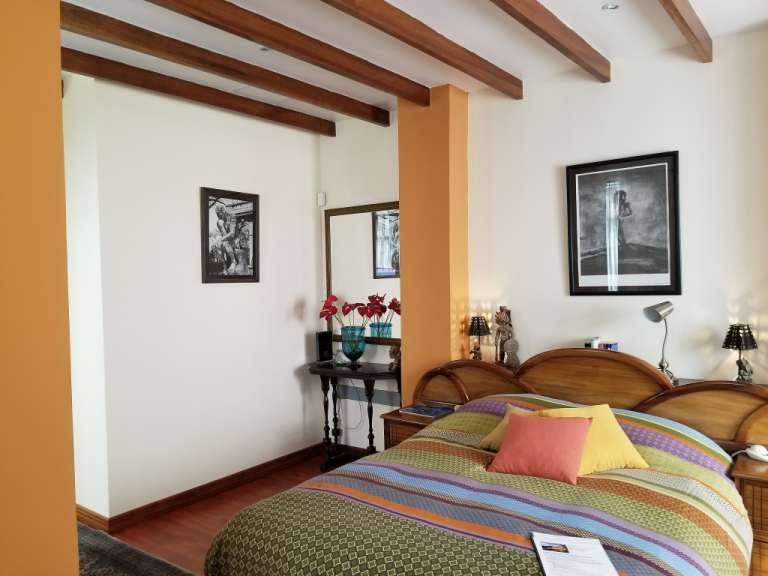 In einem Schlafzimmer mit Holzbalkendecke und orangefarbenen Säulen stehen ein Bett mit farbenfroher Bettwäsche und ein kleiner Nachttisch neben einem Spiegel.