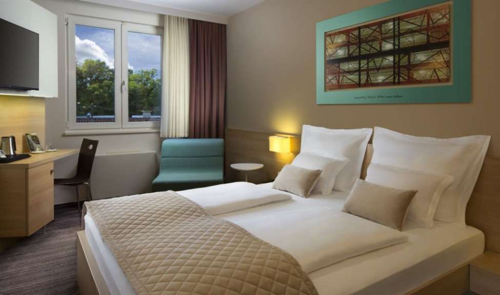 Das Bild zeigt ein Doppelzimmer mit zwei Betten, einem Schreibtisch und einem Fenster in einem Hotel.
