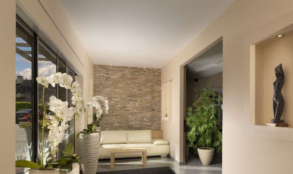Ein heller, modern gestalteter Eingangsbereich mit weißen Orchideen, einer cremefarbenen Couch vor einer Steinwand und einer Büste in einer Nische.