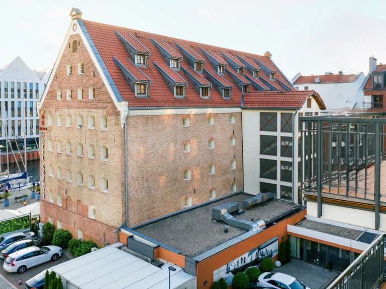 Das Bild zeigt ein Backsteingebäude mit roten Ziegeldächern und mehreren Fenstern, das von einem modernen Gebäude mit Glasfassade und einem Balkon umgeben ist.