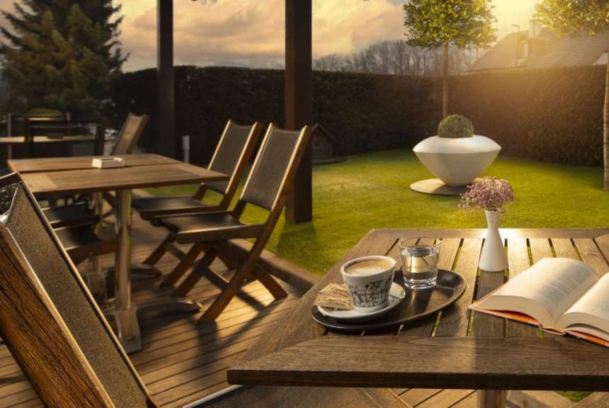 Ein offenes Buch, eine Kaffeetasse und ein Glas Wasser stehen auf einem Holztisch im Freien mit Blick auf einen Garten.