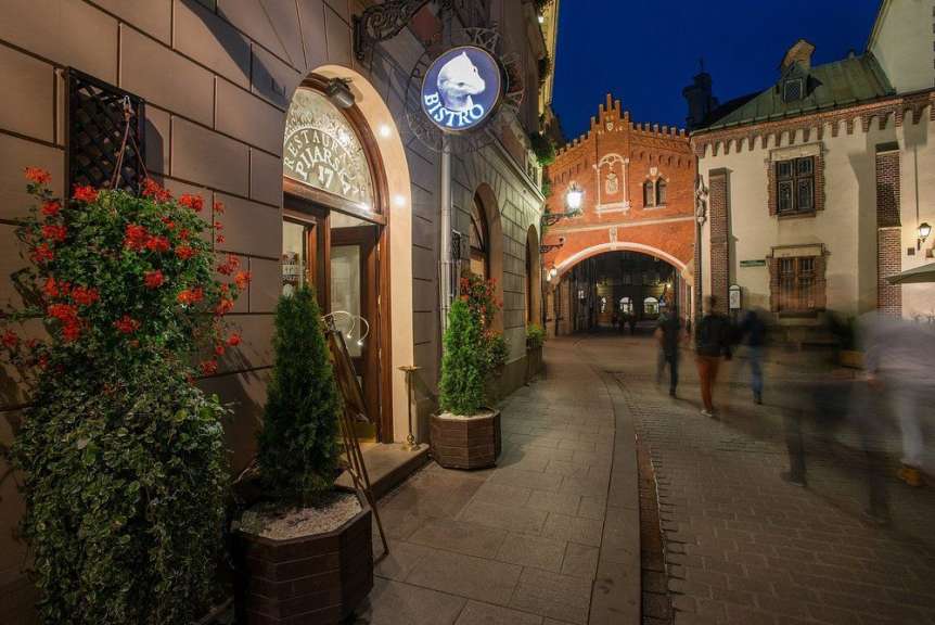 Eine Gasse mit gepflastertem Weg und Gebäuden rechts sowie Blumen und Bäumen vor einem Restaurant links führt zu einem Torbogen.