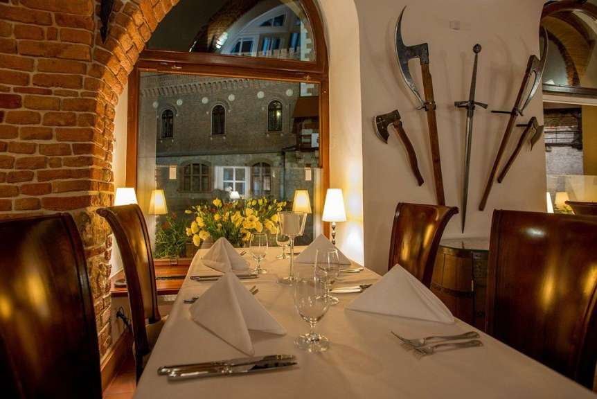 An einem mit weißem Tischtuch gedeckten Tisch in einem rustikalen Restaurant stehen schön arrangierte weiße Servietten und Besteck bereit, während durch ein Fenster die nächtliche Stadtkulisse zu sehen ist.