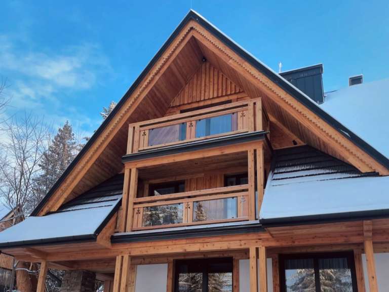 Ein hölzernes Haus mit einem spitzen Dach und schneebedeckten Giebeln steht unter einem blauen Himmel.