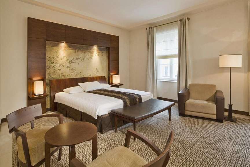 Ein geschmackvoll eingerichtetes Hotelzimmer mit zwei Betten, einem Sessel, einem Tisch und einem Fenster, das von Vorhängen eingerahmt wird.