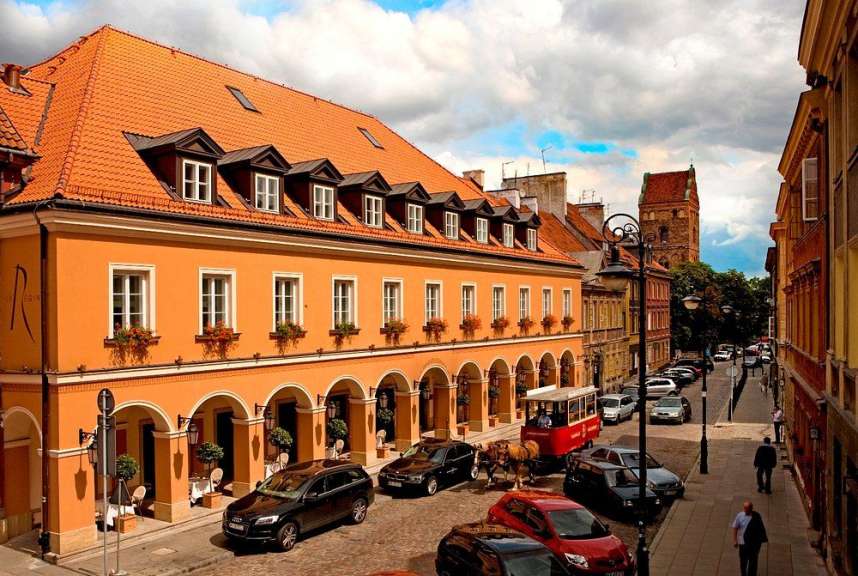 Ein Pferdefuhrwerk fährt auf einer gepflasterten Straße an einem orangenen Gebäude mit Torbögen entlang, während auf beiden Straßenseiten Autos parken und Gebäude mit roten Ziegeldächern zu sehen sind.