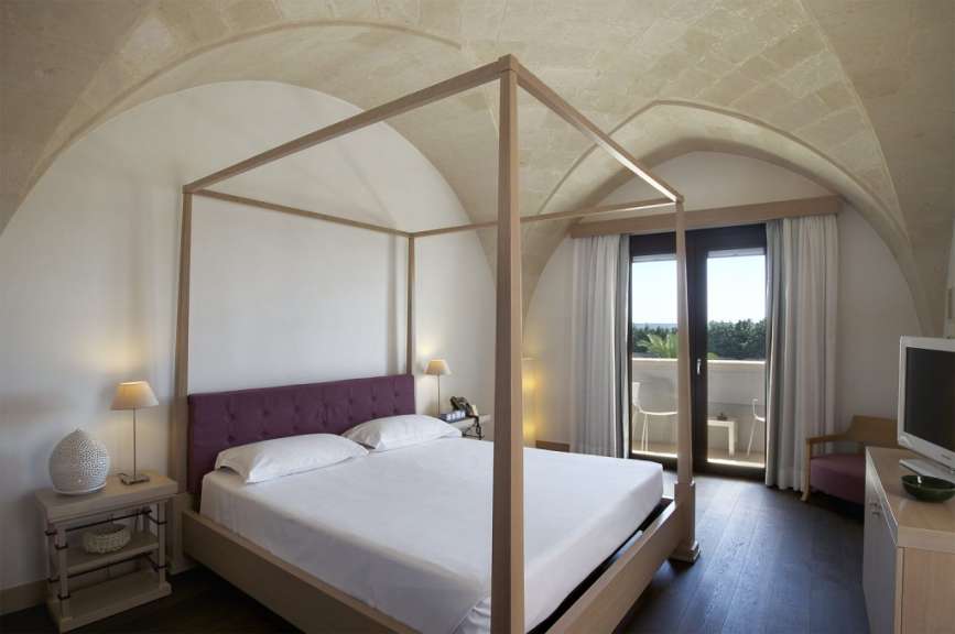 Ein Schlafzimmer mit einem Himmelbett, einem Nachttisch mit Lampe und einem Fenster mit Blick auf einen Balkon.