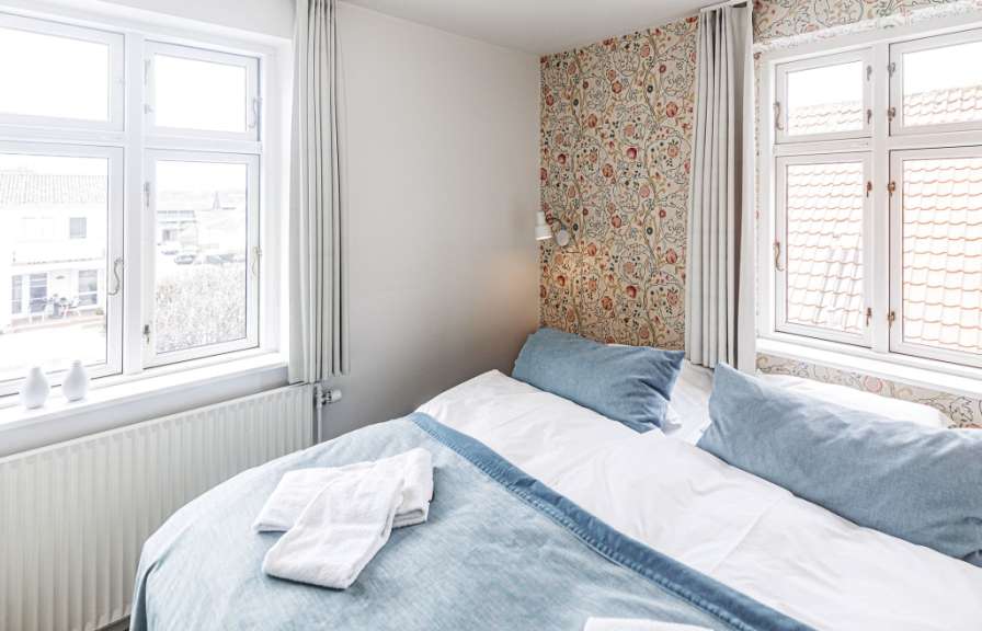 Ein Bett mit blauen Kissen und gefalteten weißen Handtüchern steht vor einem Fenster mit einem Blumenmuster auf der Tapete.