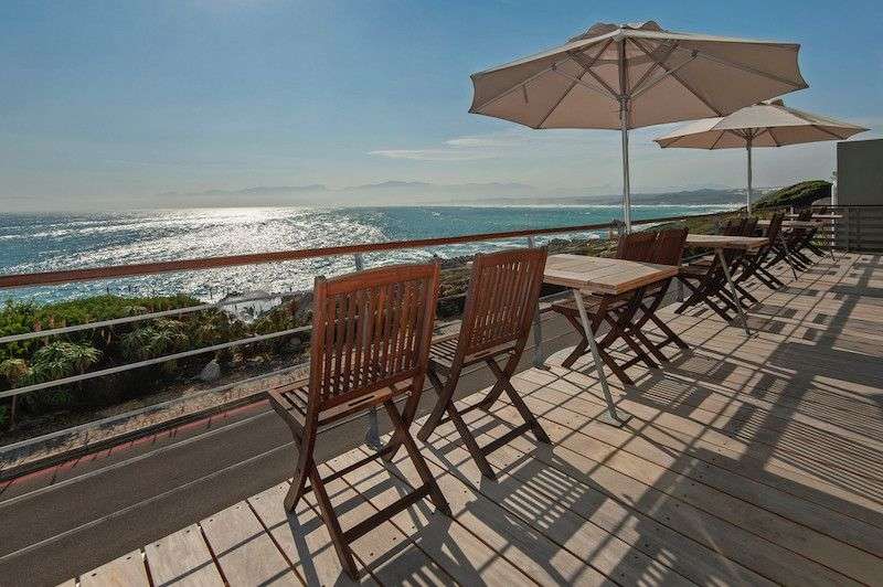 Auf einer hölzernen Terrasse mit Blick auf das Meer stehen Tische und Stühle unter Sonnenschirmen.