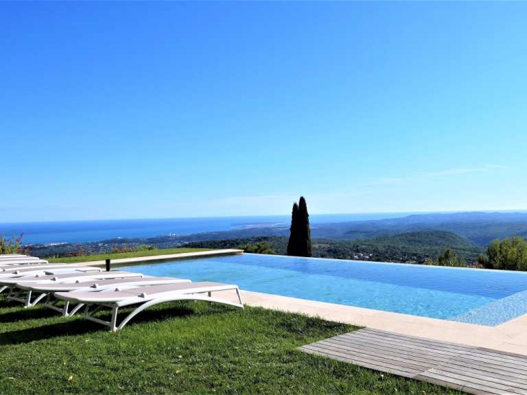 Ein Infinity-Pool mit Liegestühlen davor und einem weiten Blick auf das Meer und die umliegenden Berge im Hintergrund.