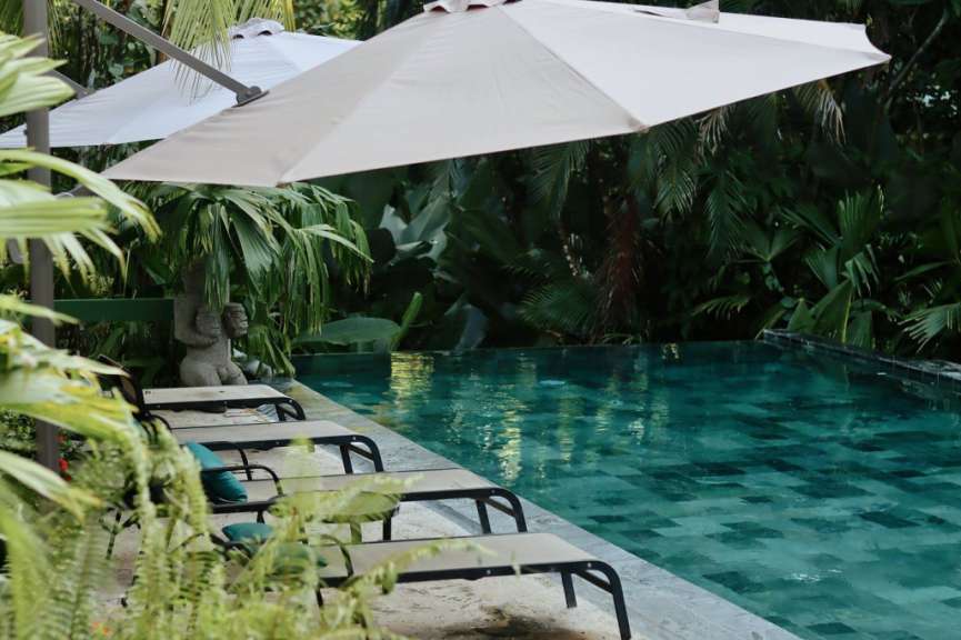 Ein Pool mit türkisfarbenem Wasser und eine Reihe von Sonnenliegen stehen unter einem großen Sonnenschirm neben üppiger Vegetation.
