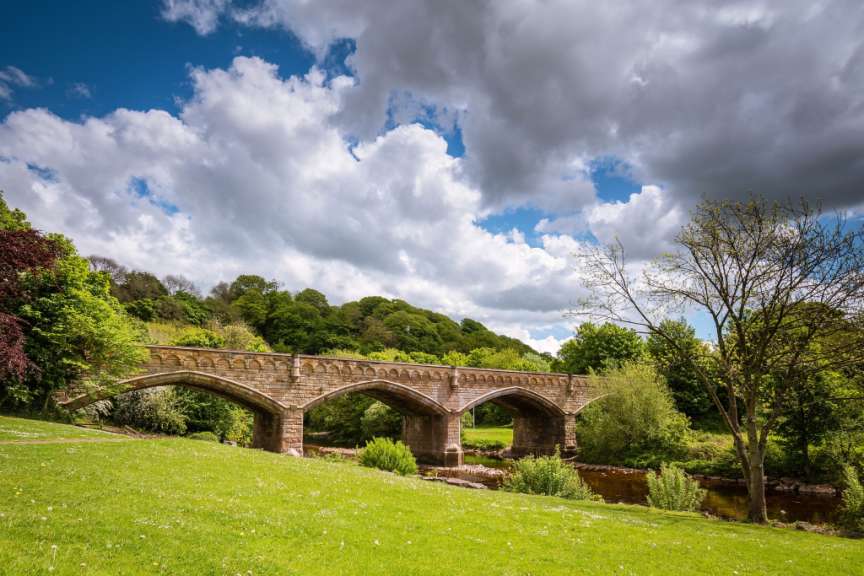 Ein steinerner Bogenbrücke überspannt einen Fluss, umgeben von üppig grüner Vegetation und einem bewölkten Himmel.