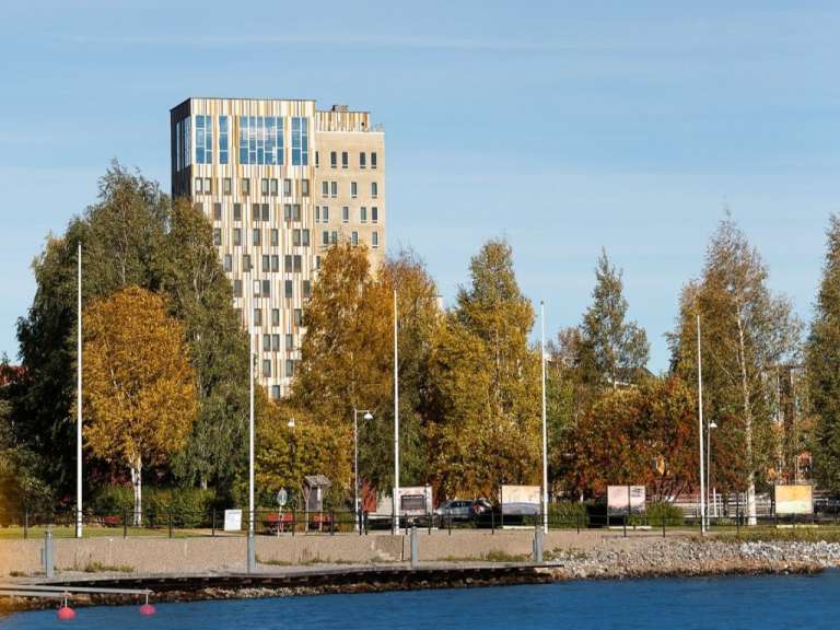 Ein Moderner Hochhaus mit goldenen und weißen Fassaden steht hinter einer Reihe von Bäumen in Herbstfarben, mit einem blauen Wasserufer und klarer Himmelskulisse.