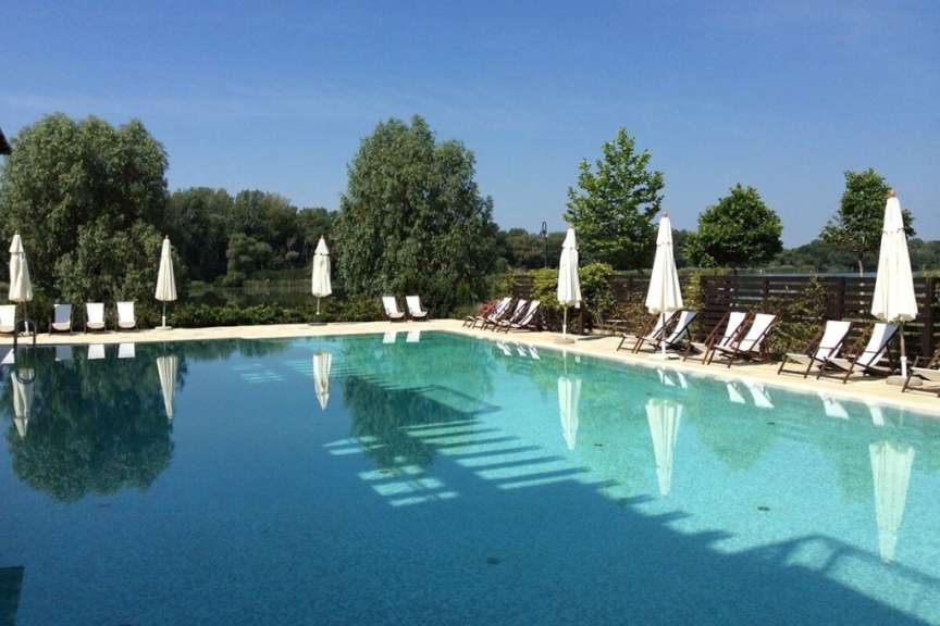 Ein Schwimmbecken mit Liegestühlen und Sonnenschirmen am Ufer eines Sees mit Bäumen im Hintergrund.