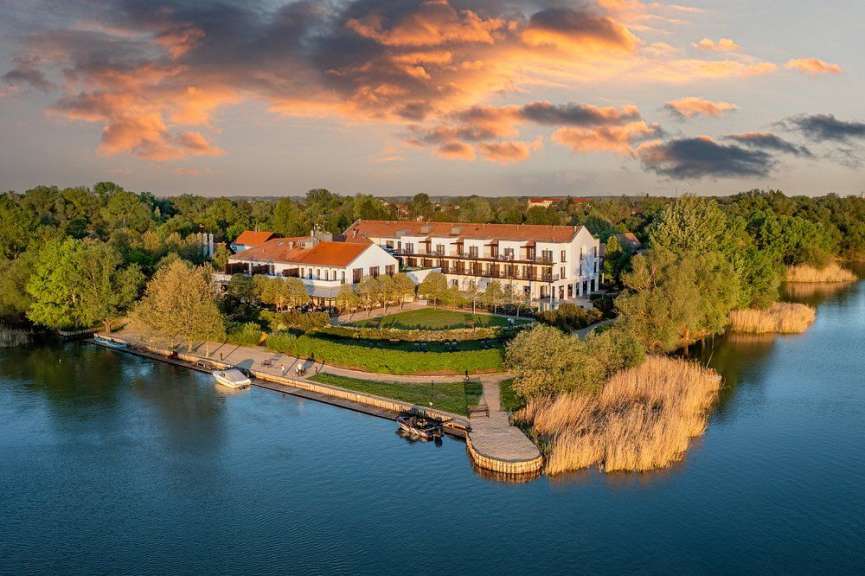 Ein Hotel mit weißen Fassaden und roten Dächern liegt an einem See inmitten grüner Bäume und trockener Schilfbestände bei Sonnenuntergang.
